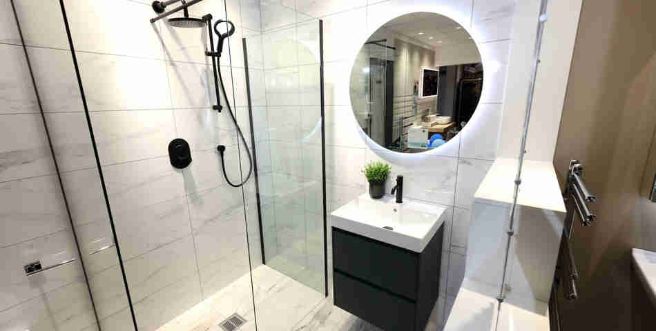 A modern style bathroom featuring pale marble tiles, a walk in shower, sink on top of a black cabinet and a round mirror on the wall