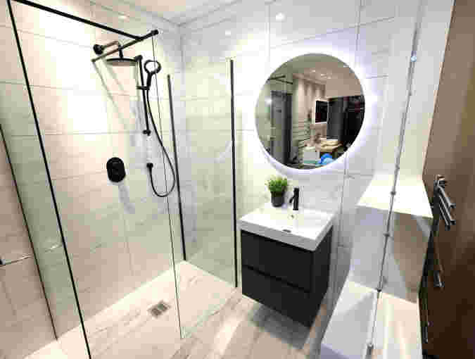 A modern style bathroom featuring pale marble tiles, a walk in shower, sink on top of a black cabinet and a round mirror on the wall