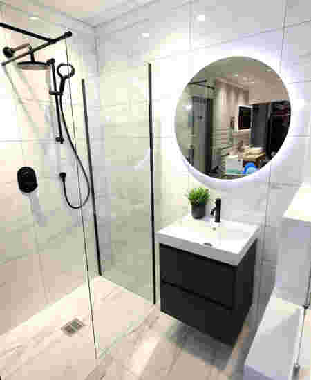 A modern style bathroom featuring pale marble tiles, a walk in shower, sink on top of a black cabinet and a round mirror on the wall